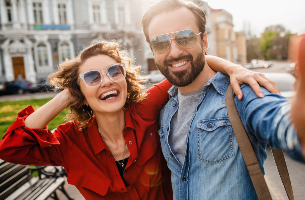 Young happy couple taking a selfie while wearing sunglasses to protect vision from dry eye factors.