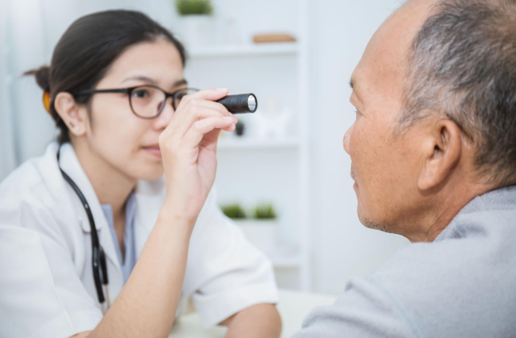 A senior man facing his optometrist as she shines a small flashlight into his eye
