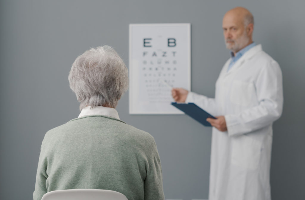 View from behind of an older adult woman's head as she sits in a chair and looks ahead. Her eye doctor is pointing to an eye chart on the wall.