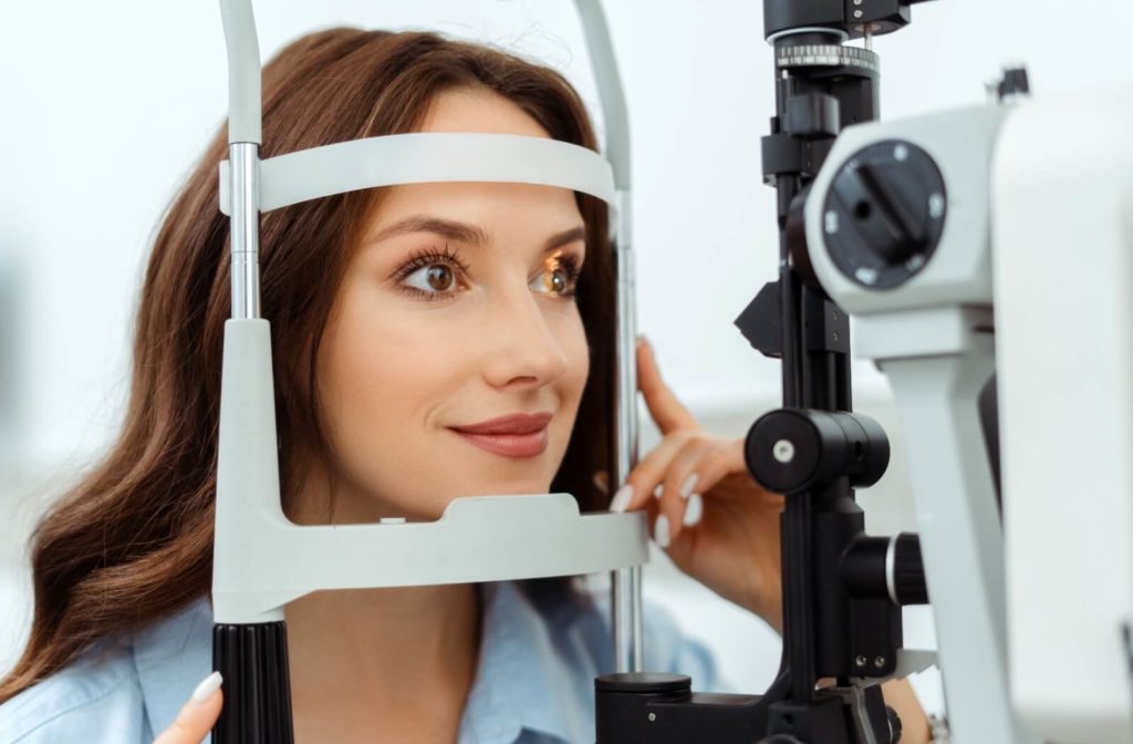 A smiling patient sits in an optometrist's office, resting their chin in front of a slit lamp during their routine comprehensive eye exam.