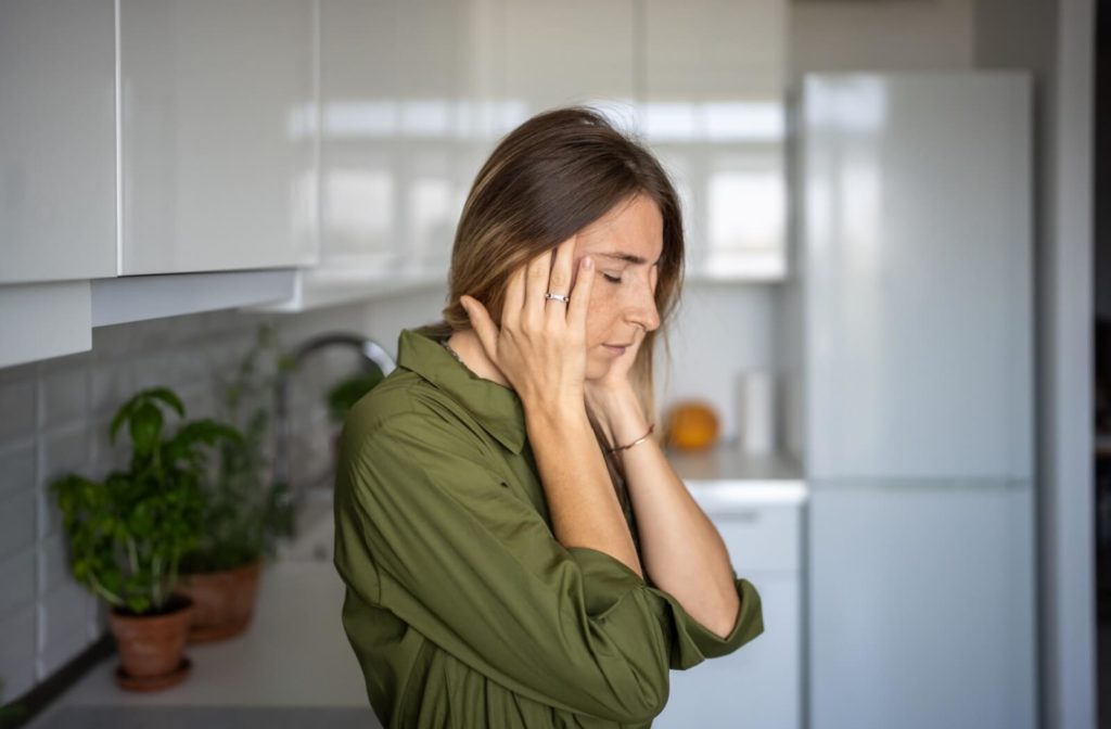 A person in a green shirt stands in the kitchen, hands pressed to their temples, visibly uncomfortable from a headache triggered by dry eye.