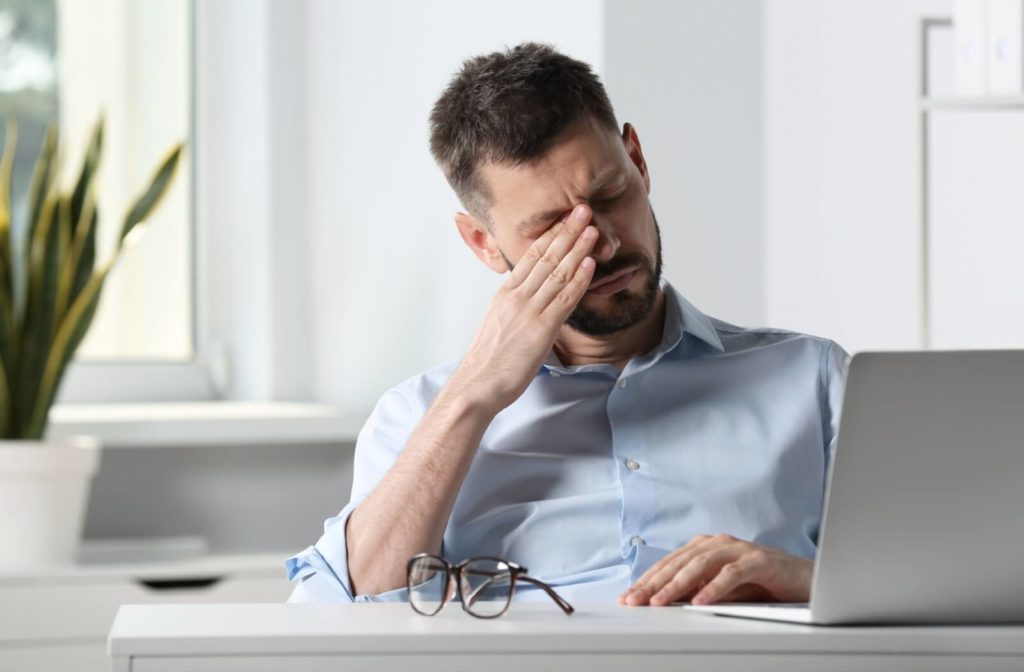 Seated at a computer, a person rubs the bridge of their nose with their right hand, seeking relief from a headache caused by dry eyes.